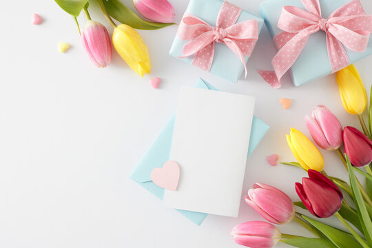 Mother's Day concept. Top view photo of envelope with card gift boxes with bows colorful hearts and bouquets of flowers yellow pink tulips on white background with empty space