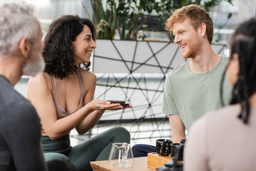 happy middle eastern woman holding compressed puer tea near redhead man in yoga studio.