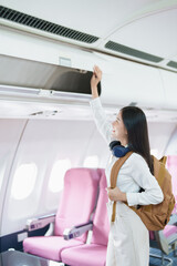 Young asian attractive woman travel by airplane, Passenger wearing headphone putting hand baggage in lockers above seats of plane