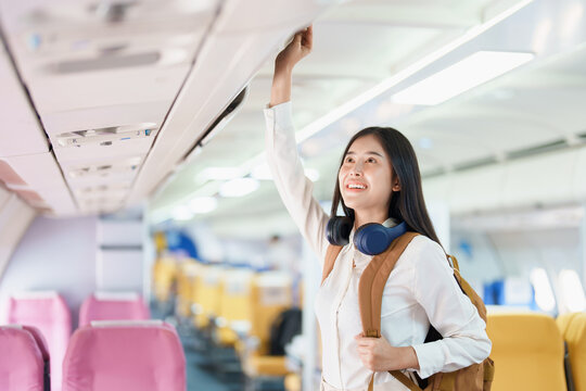 Young asian attractive woman travel by airplane, Passenger wearing headphone putting hand baggage in lockers above seats of plane