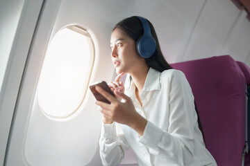 Young Asian attractive business woman passenger sitting on business class luxury plane while wearing wireless headphone and using smart phone mobile for relax during the flight