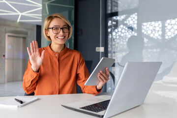 A young female student sitting in an office, co-working space, library at a table with a laptop and using a tablet to study. Smiling, she looks at the camera. Talks on a video call, waves his hand.