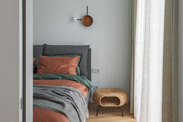 A view through an open door into a bedroom interior with rust color linen and cushions on a bed, grey blanket, rattan bedside table and metal wall light.