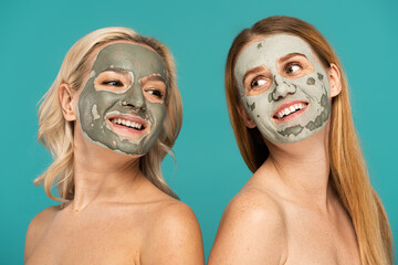 cheerful redhead and blonde women with clay mask on faces posing isolated on turquoise.