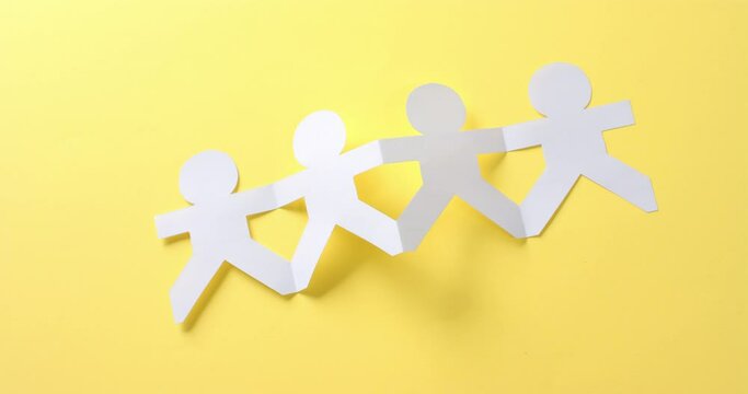 Close up of people holding hands made of white paper on yellow background with copy space