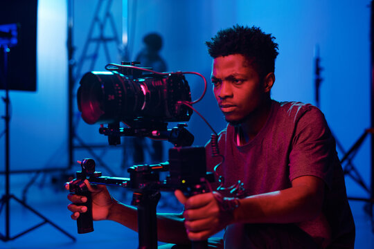 Young African American camera operator shooting on professional camera in dark studio