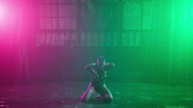 Cinematic video of young gorgeous professional female dancer in wet bodysuit performing erotic seductive choreography on wet floor in urban garage with dramatic rain and lighting on cyber background
