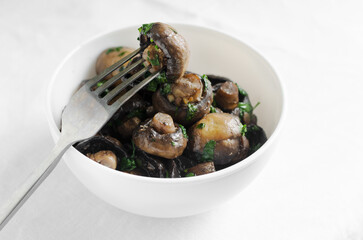 Fried champignons with garlic and parsley in a white bowl with a fork with a mushroom. The concept of vegetarian food. Horizontal orientation. Selective focus.