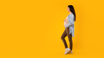 Fototapeta na wymiar Pregnant lady embracing her belly, looking at free space and smiling, yellow background, panorama, side view