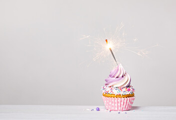 Purple and pink cupcake with sprinkles and lit sparkler on a white grey background. - 588462282