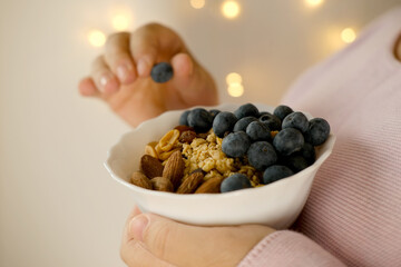 close-up female hand takes berries on blurred background with bokeh, healthy food white cup, oatmeal, blueberries, nuts, almonds, peanuts , raisins, dried fruits, raw food diet, vegetarian food