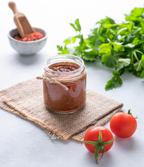 Hot sauce adjika. Homemade appetizer with pepper and tomatoes in a jar on light background with fresh herb and vegetables.