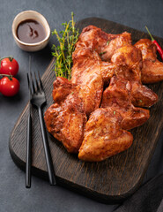 Spicy grilled chicken wings BBQ with sauce on a wooden plate with tomato and herb on a dark background of slate or concrete.