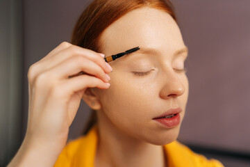 Close-up face of beautiful redhead young woman applying light shadows to eyebrows with make-up brush. Portrait of ginger female combing eyebrows with makeup brush. Concept steps of make-up applying.