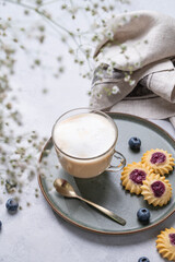 Cappuccino or latte with milk foam in a cup with homemade berry cookies and blueberries on a light background with gypsophila branches. Concept spring morning breakfast.
