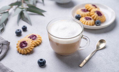 Cappuccino or latte with milk foam in a cup with  homemade berry cookies and blueberries on a light background with eucalyptus branches with shadow.