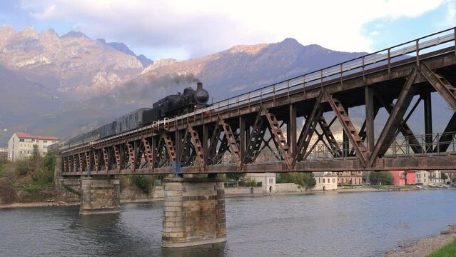 Ancient steam train crossing an ancient iron bridge over a river with mountains in the background. steam locomotive