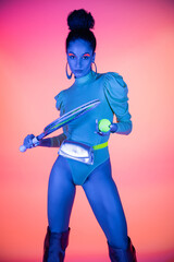 Fototapeta premium Trendy african american woman in bodysuit holding tennis racket and ball on pink background with neon light.