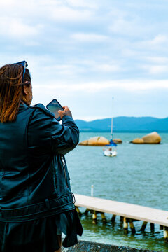 Mulher turista tirando foto de praia com veleiro e pedras ao fundo Coqueiros, Florian&oacute;polis, Santa Catarina, Brasil