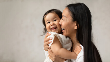 Joyful Asian Mom Hugging Baby Daughter Over Gray Wall Indoor
