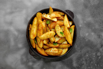 Roasted potatoes with herbs and spices. Baked potato wedges in frying pan on dark stone background. Top view with copy space.