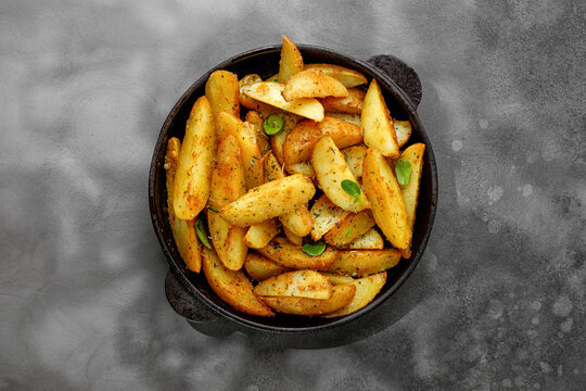Roasted potatoes with herbs and spices. Baked potato wedges in frying pan on dark stone background. Top view with copy space.