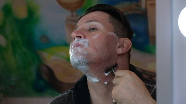 A man shaves with an old style razor with a replaceable blade and a foam shaving brush. Traumatic shave. The old way of skin and beard care. Bristle haircut. Soaped face close-up