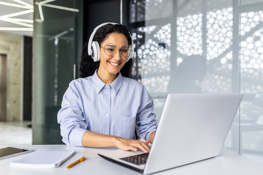 Young beautiful Indian female programmer working inside office with laptop, woman in headphones listening to audio podcast and online course, typing on keyboard and reaching out, female engineer .