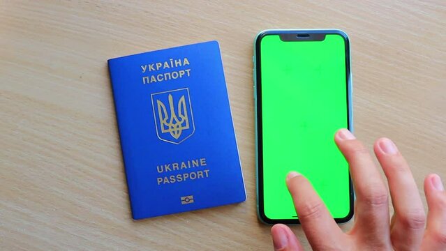 Woman's hand scrolling green screen browsing chroma key reading social media close up with Ukrainian passport and smartphone on the table