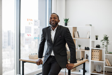 Smiling multiethnic company leader resting on edge of writing desk while posing in comfortable workplace. Joyful entrepreneur in business suit taking break from managing performance of organization.