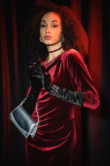 Young african american woman in velvet red dress and gloves holding handbag near burgundy drapery.