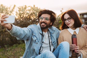 Cheerful young diverse man and lady in casual and glasses with thermos take selfie on smartphone