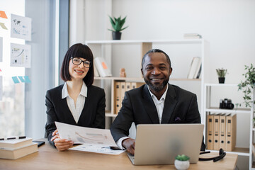 Smiling caucasian woman and african american man in formal clothes posing at writing desk with modern laptop and documents. Efficient colleagues having break from coworking process in office.