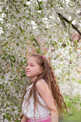 Adorable 9 years old girl with long curly hair surrounded by plenty branches blossoming of white spring flowers. Portrait of young girl
