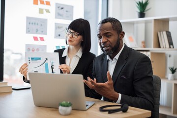 Confident multicultural man communicating online via laptop while attractive caucasian woman demonstrating increase graphs. Efficient business partners discussing project over during video call.