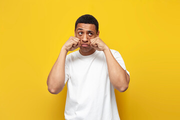 young sad guy african american in white t-shirt is crying on yellow isolated background, unhappy...