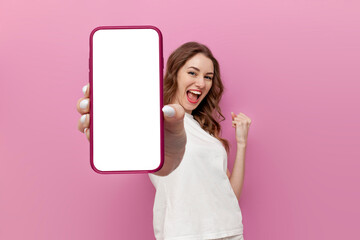 young cute woman in white t-shirt shows blank smartphone screen and rejoices in victory on pink...