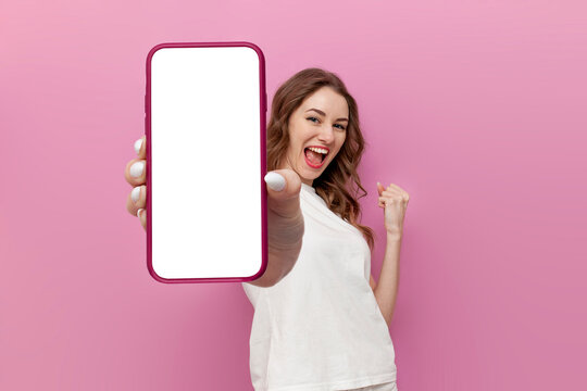young cute woman in white t-shirt shows blank smartphone screen and rejoices in victory on pink isolated background
