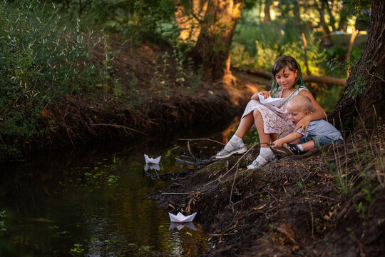 Girl and boy are sitting on the bank of river, launching white paper origami boat into the water.