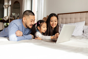 asian family use laptop at home on bed, korean dad mom and little daughter look at computer screen and study online