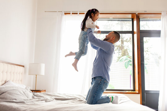 small active Asian girl plays with her dad at home, energetic child jumps and dances on the bed with parent