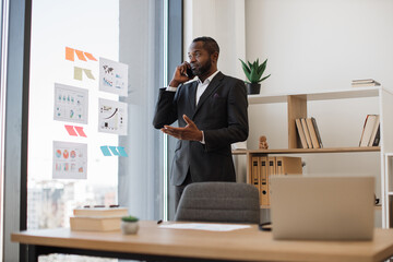 Multiethnic businessperson in formal outfit having conversation on cell phone while standing out against office background. Confident male employer thinking over speaker's words in cozy workplace.