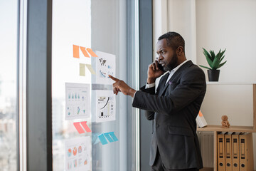 Focused multiracial businessman in smart suit receiving phone call while standing near glass panel with graphs and charts in corner office. Senior executive listening to worker's speech over mobile.