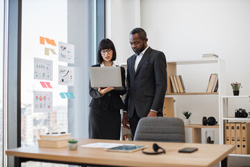 Young caucasian businesswoman displaying information on laptop to multiethnic male colleague standing in modern office. Efficient coworker presenting her ideas of new corporate strategy.