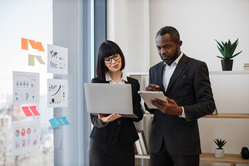 Caucasian woman and multiracial man in business clothes holding electronic devices while communicating in modern workplace. Professional coworkers being involved in teamwork using laptop and tablet.