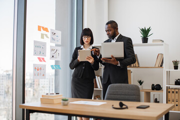 Confident entrepreneurs working together in office with help of modern handheld devices. Multiethnic businessman with caucasian female partner discussing market growth looking on graphic materials.