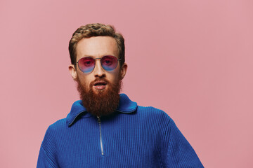 Portrait of a redheaded man wearing sunglasses smiling and dancing, listening to music on a pink background. Hipster with a beard, happiness finger pointing.