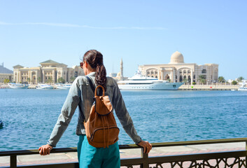 Fototapeta premium A young woman tourist with a backpack looks at the administrative region of the emirate of Sharjah with the port and ships.