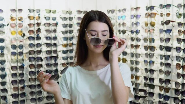Portrait of cheerful hesitating woman in optician store, making decision, holding stylish sunglasses, choosing what she should buy. Girl on a background of showcase with different models of glasses
