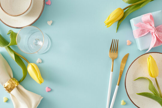 Mother's Day concept. Top view photo of gift box plate cutlery knife fork fabric napkin cup and glasses yellow tulips and colorful hearts on pastel blue background with empty space in the middle